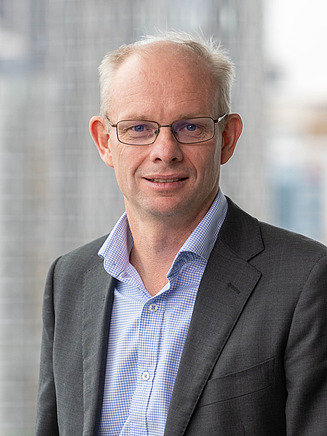 Scott Morris, representing the STRABAG Australia Management and Leadership Team. The image shows a business professional standing in a modern office environment near floor-to-ceiling windows, wearing business attire.