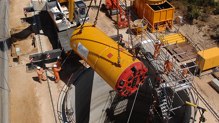 A large tunnel boring machine component is being lowered into a circular construction shaft by a heavy-duty crane. The image shows an active infrastructure construction site with workers in safety gear, support vehicles, machinery, equipment, and temporary site facilities surrounding the excavation area.
