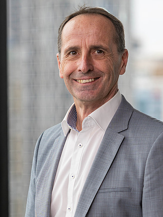 Professional business portrait of Herbert Krutina, a STRABAG Australia leadership representative standing in a modern office environment. The image shows him wearing a grey blazer and white shirt, positioned in front of large floor-to-ceiling windows with natural daylight and