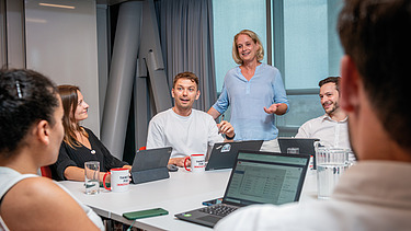 Employees participate in a project meeting in a modern conference room, with laptops and tablets placed on the table. One team member presents to colleagues while others listen and collaborate during a discussion focused on project planning, business operations, and teamwork.