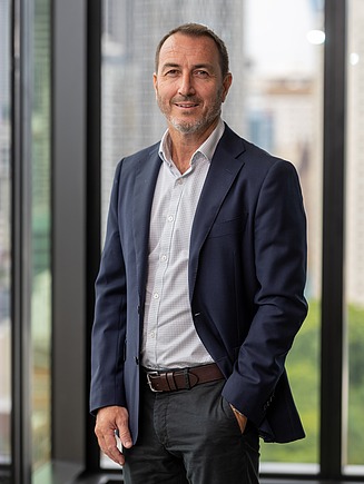 Professional portrait of Gary Georgiou, representing the STRABAG Australia Management and Leadership Team. The image shows him standing in a modern office environment near floor-to-ceiling windows, wearing business attire.