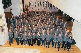 Large group of Georgiou Group employees gathered for a team photograph inside a modern office building. Team members wearing matching company-branded grey shirts are arranged across a staircase and open lobby area, with contemporary architectural features visible in the background