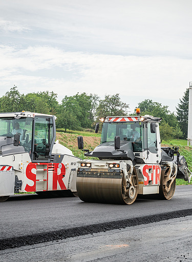 Three STRABAG road rollers compact fresh asphalt on a newly paved roadway, showcasing modern construction equipment, precision engineering, and efficient infrastructure delivery in a rural environment with industrial silos in the background.