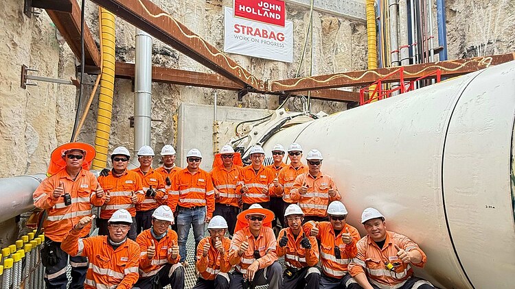 Project team members wearing high-visibility orange safety clothing and white hard hats pose inside the Belmont Desalination Plant Intake Tunnel construction site. The image shows a large tunnel pipeline, excavation support structures, and underground infrastructure, with a “Breakthrough” milestone sign in the background marking the successful completion of a key tunnelling phase