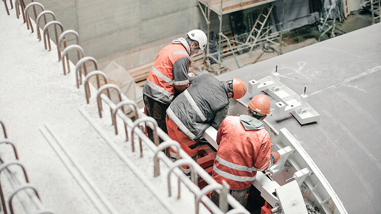 Construction workers in high-visibility safety clothing and hard hats perform assembly work on a tunnel boring machine (TBM) inside an underground tunnel construction site. 